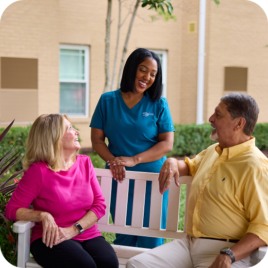 A nurse in teal scrubs smiles and talks with an older woman in a pink shirt and an older man in a yellow shirt—St.Louis Seniors enjoying friendly SeniorConcierge care together on a bench outdoors.