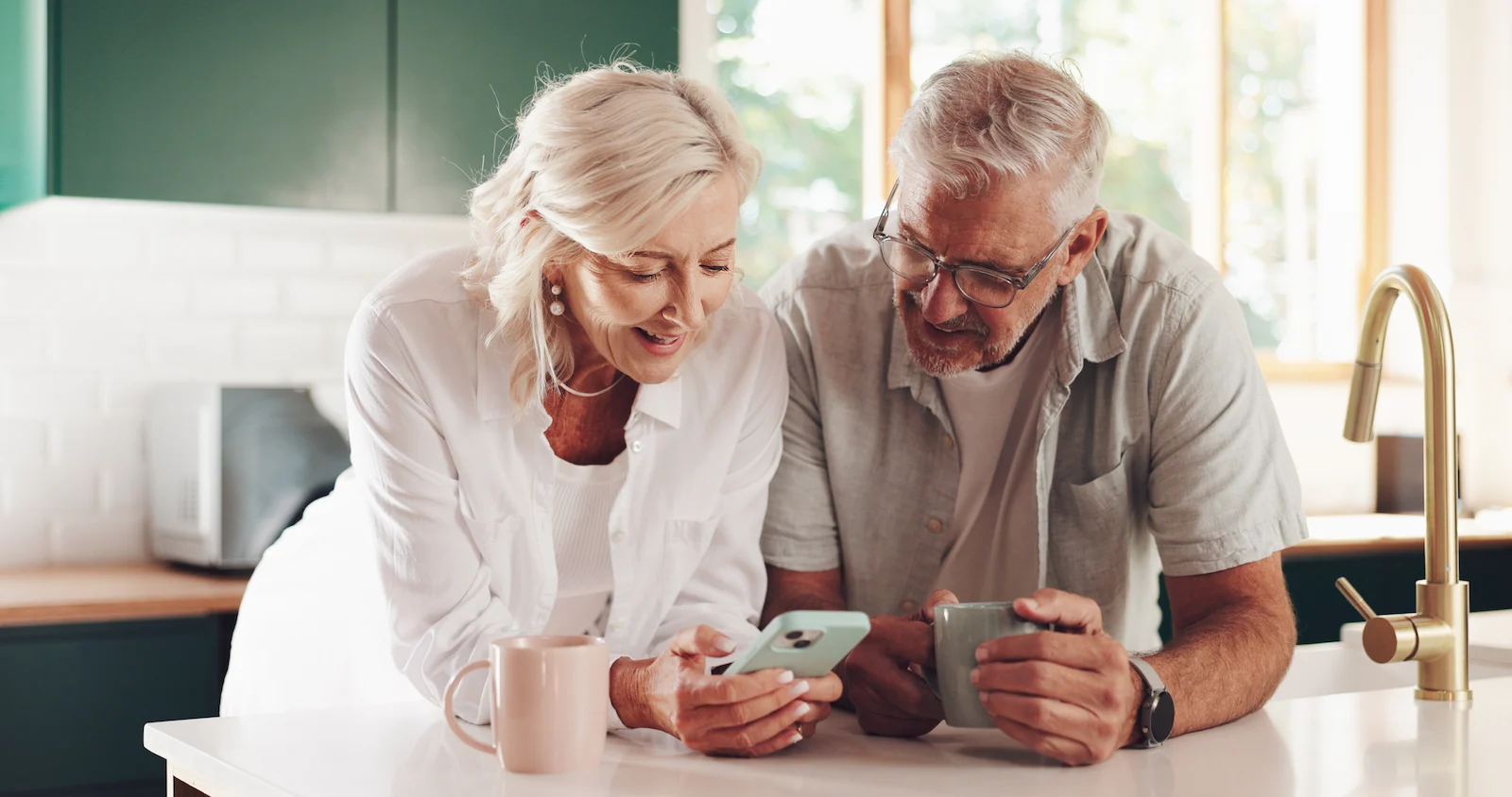 An elderly couple smiles while looking at a smartphone together in a bright kitchen. They are leaning on a counter, each holding a mug, appearing happy and relaxed.
