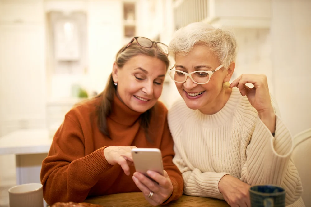 Two older women sit at a table indoors, smiling and looking at a smartphone together. One woman wears glasses and an orange sweater, the other has short gray hair, white glasses, and a cream sweater.