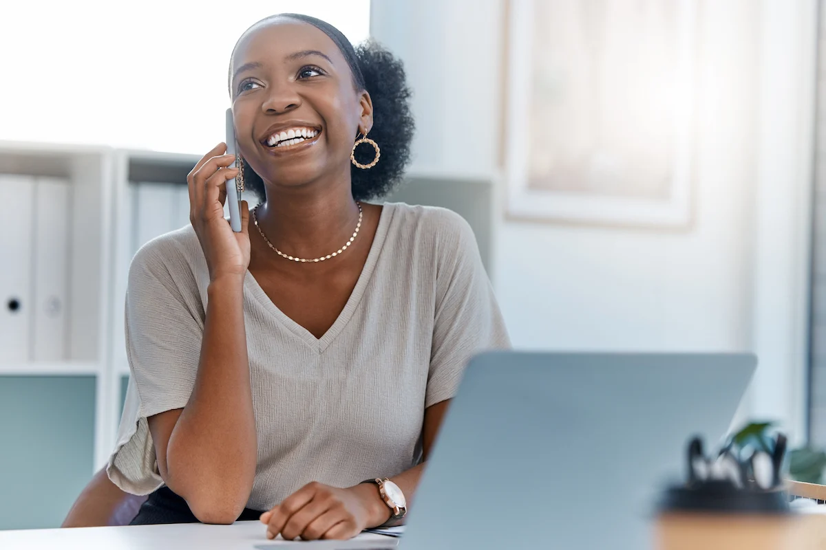 A woman smiling and talking on a smartphone while sitting at a desk with a laptop, wearing a light blouse and gold hoop earrings in a bright, modern office setting.