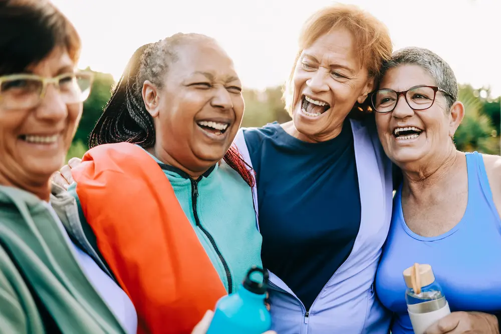 Four St.Louis Seniors in athletic clothes stand close together outdoors, smiling and laughing. They appear happy and relaxed, with one holding a water bottle. The bright, slightly blurred background adds to the joyful scene.