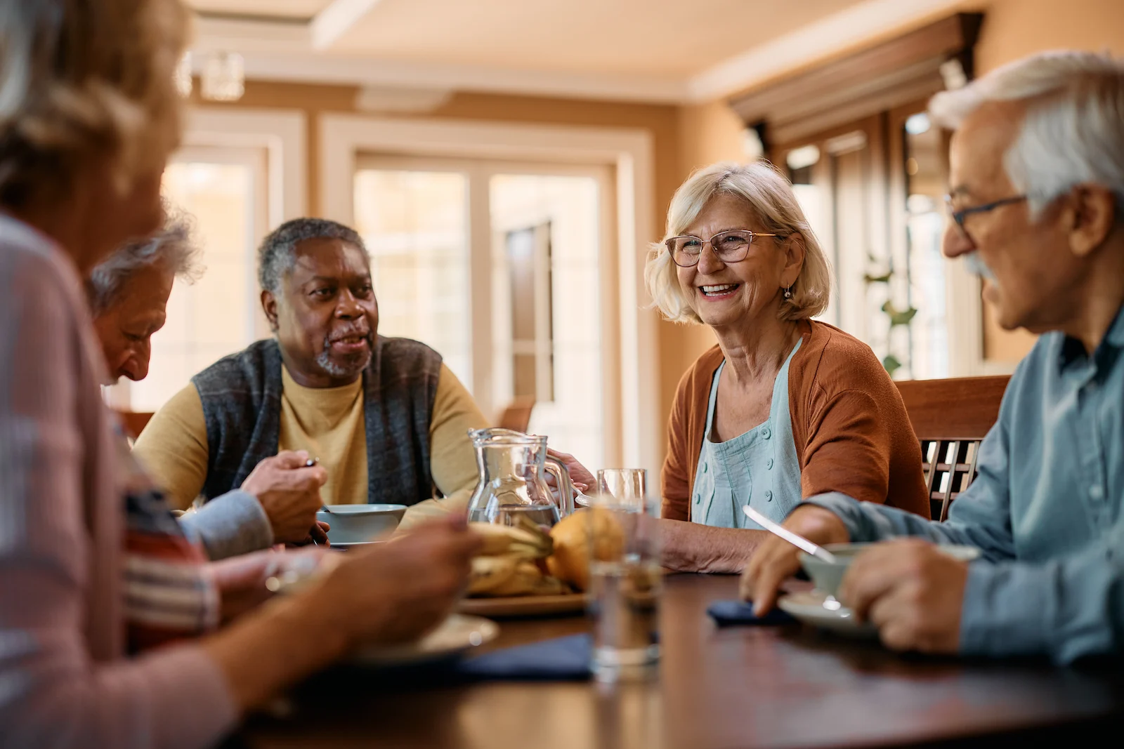 A group of older adults sit around a dining table, smiling and talking while sharing a meal in a bright, cozy room with large windows.