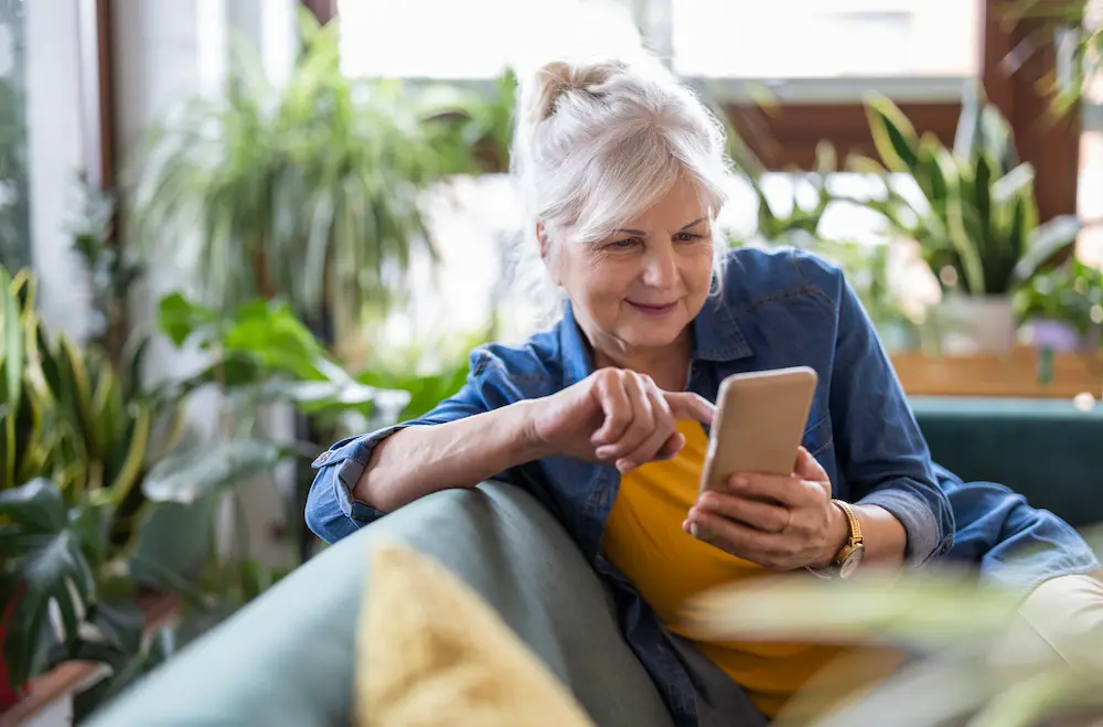 An older woman with gray hair sits on a couch in a bright room, smiling and using her smartphone. Surrounded by green plants, she appears relaxed and engaged—just the kind of day SeniorConcierge St.Louis Seniors can help you enjoy.
