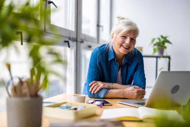 Older woman with gray hair, wearing a blue shirt, smiling while sitting at a desk with a laptop, notebook, and plants nearby. Bright, modern room with large windows and natural light.