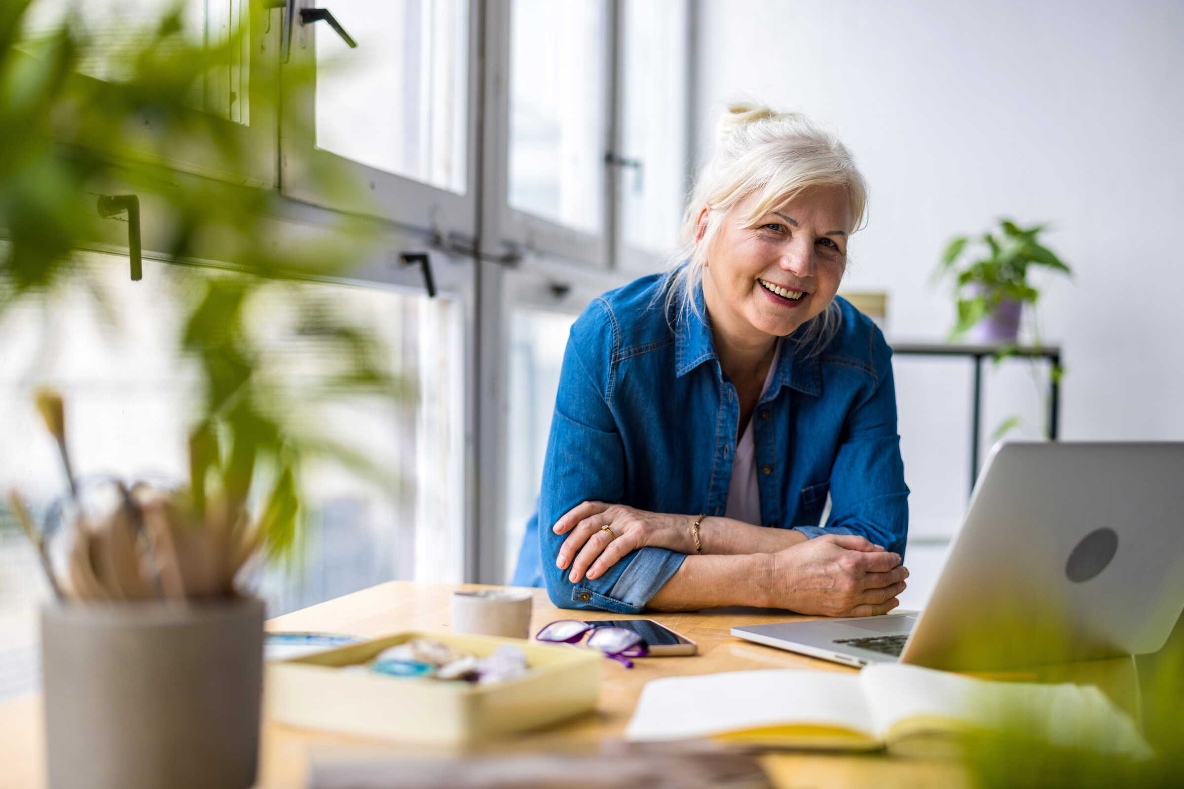 Older woman with gray hair, wearing a blue shirt, smiling while sitting at a desk with a laptop, notebook, and plants nearby. Bright, modern room with large windows and natural light.