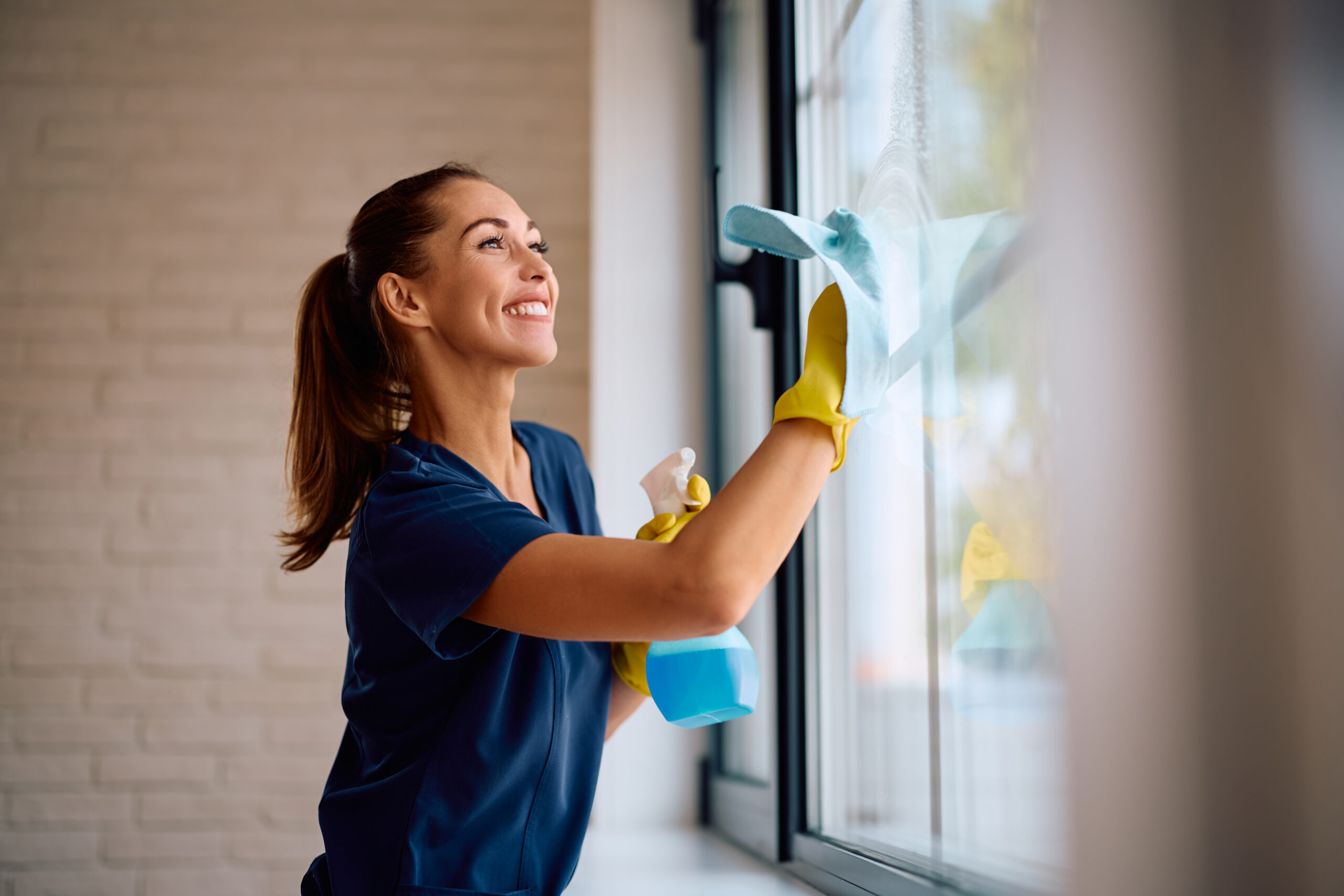 A smiling woman wearing yellow rubber gloves and a blue shirt is cleaning a window with a blue cleaning cloth and a spray bottle. She appears to be enjoying the task in a bright, modern room.