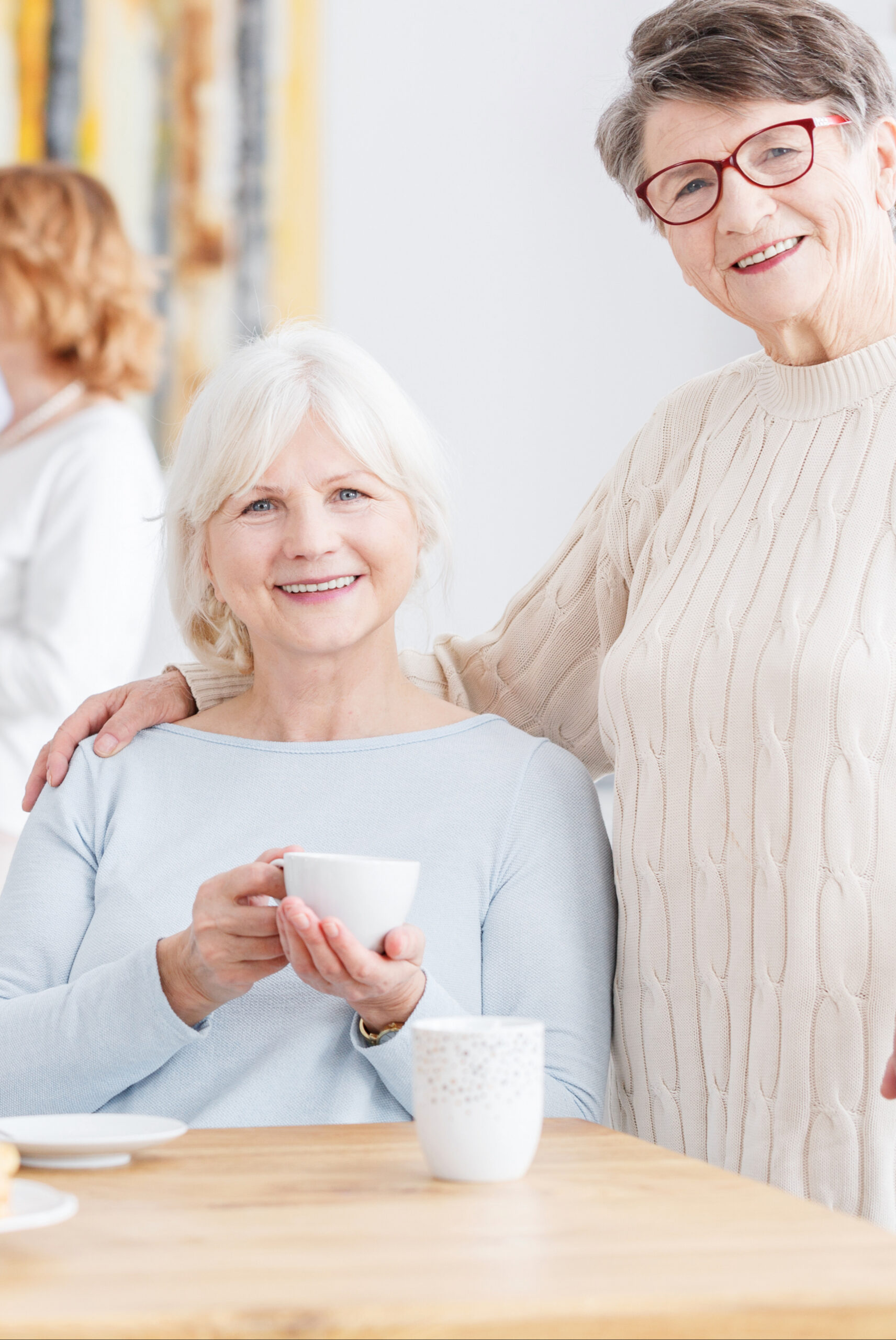 Two older women smiling together indoors; one is sitting and holding a cup, while the other stands beside her with a hand on her shoulder. Another woman appears blurred in the background.